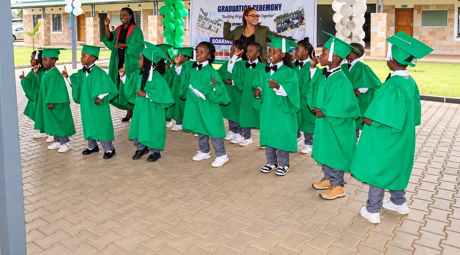 Graduates in green caps and gowns celebrating with their teachers in front of the graduation ceremony banner